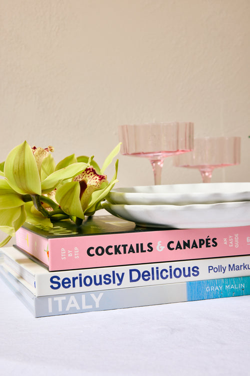 Stack of cookbooks titled 'Cocktails & Canapes', 'Seriously Delicious', and 'Italy' on a surface with a decorative plate and flowers.