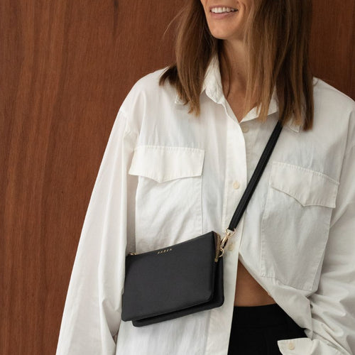 Woman wearing a white shirt and black pants, holding a black crossbody bag against a wooden background