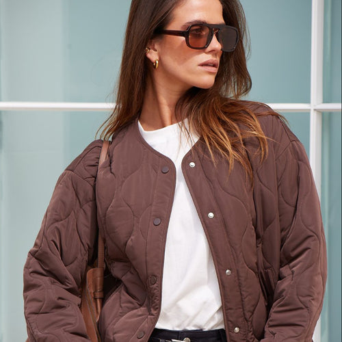 Woman wearing a brown quilted jacket and sunglasses against a light blue wall.