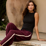 Woman sitting on a wooden step wearing a black sleeveless top and burgundy pants with white stripes.