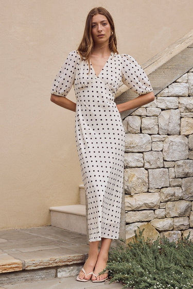 Woman in a white dress with black polka dots standing on a stone path next to a stone wall.
