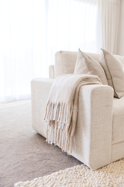 A close-up of a light beige upholstered armchair in a bright, minimalist room, styled with matching beige throw pillows and a fringed cream throw blanket on the seat.