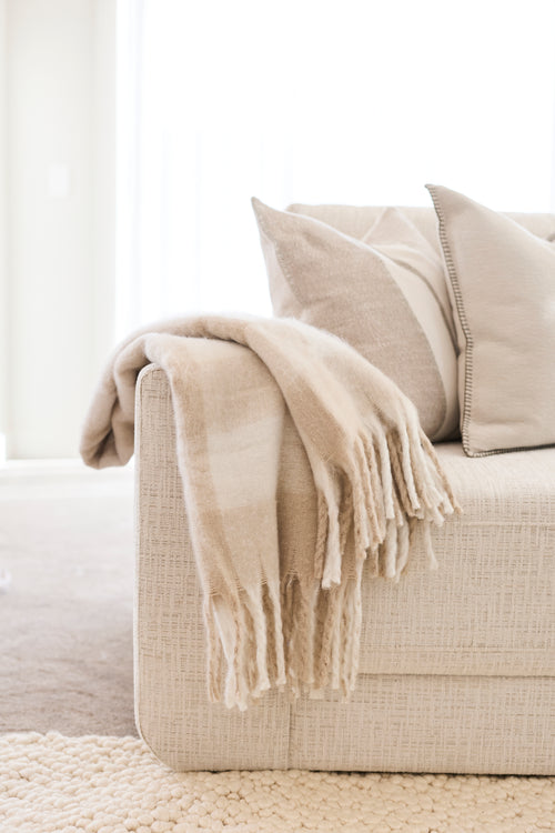 A cozy beige throw blanket with fringe is draped over the arm of a light beige upholstered sofa, alongside matching pillows in a bright, minimalist room.