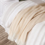 A close-up of a soft cream throw blanket draped over white bedding on a bed, with a textured taupe rug visible at the bottom.