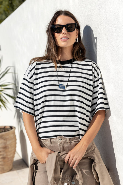 A woman wearing a black and white horizontally striped short-sleeve t-shirt, beige cargo pants, and black sunglasses, leaning against a white stucco wall outdoors.