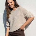 A woman posing outdoors against a white wall, wearing a beige knit sweater and a dark brown midi skirt, holding a small gray handbag.