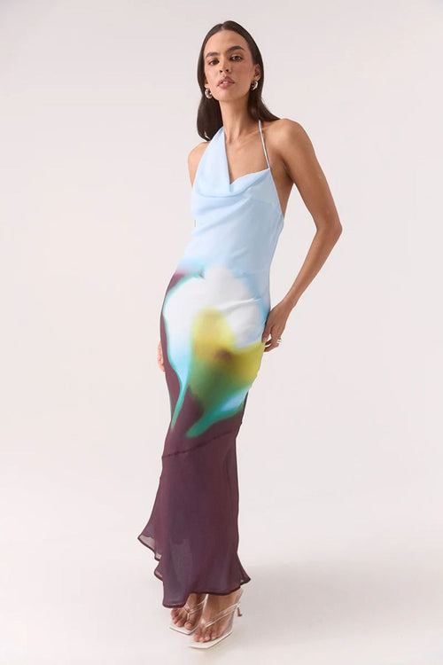 Model wears halter-neck maxi dress with blurred azure, green, and plum abstract floral print, asymmetric hem, and a softly draped neckline.