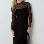 Woman wearing a black dress with sheer sleeves against a white brick wall.