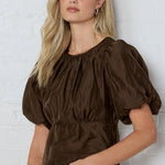 Woman wearing a brown blouse against a white brick wall