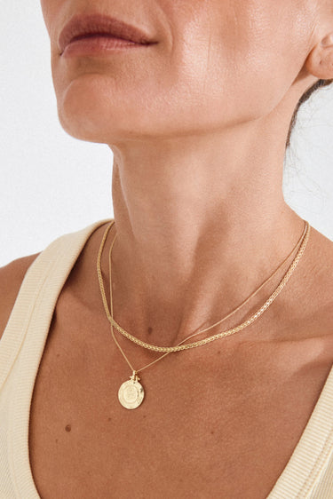 Close-up of a person wearing a gold necklace with a coin pendant on a plain background