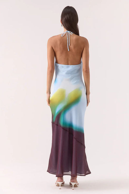Model wears halter-neck maxi dress with blurred azure, green, and plum abstract floral print, asymmetric hem, and a softly draped neckline.