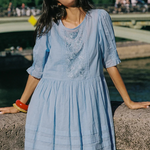 A woman wearing a light blue cotton dress with embroidered details on the chest, leaning on a stone bridge with a river and green trees in the background.