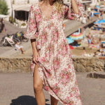 A woman wearing a long-sleeve, V-neck maxi dress with a pink and green floral print on a cream background, standing outdoors on a sunny day with a beach town background.