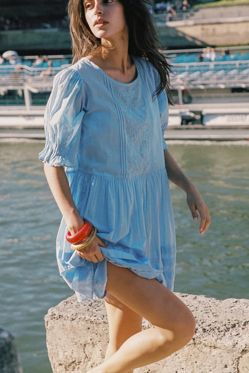 A woman wearing a light blue cotton puff-sleeve dress stands on a stone ledge by the water, holding a red and orange striped towel. The background shows a blurred outdoor harbor or marina scene.