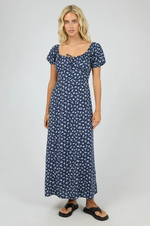 A woman modeling a navy blue maxi dress with a small white floral print, puff sleeves, and a scoop neckline with a tie detail at the front, set against a plain light gray studio background.