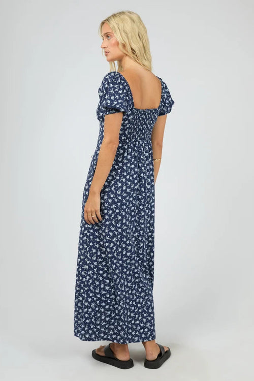 A woman modeling a navy blue maxi dress with a white floral pattern, featuring puff sleeves and a smocked bodice, shown from a three-quarters back view against a light gray studio background.