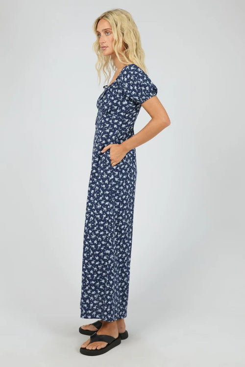 A woman modeling a navy blue maxi dress with a white floral print, standing in profile against a light gray studio background.