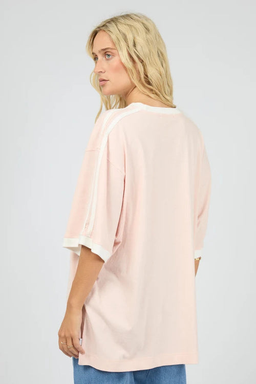 A woman wearing an oversized light pink t-shirt with white contrast trim on the sleeves and collar, shown from a three-quarter back view against a light gray studio background.