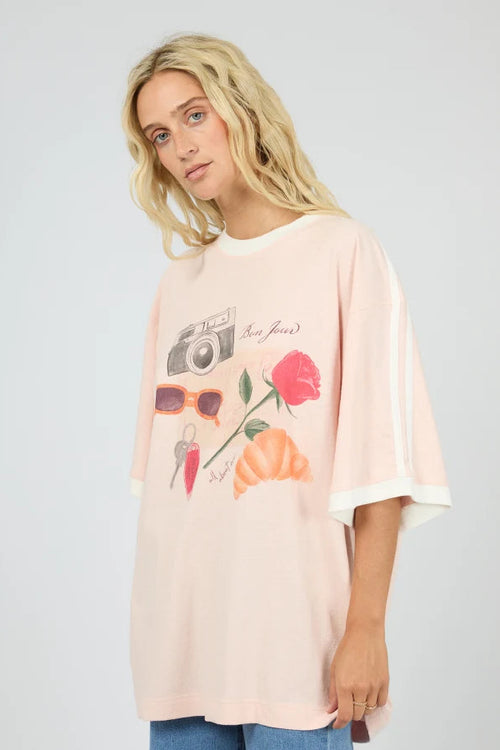 A woman wearing a light pink oversized graphic T-shirt with white sleeve stripes and a colorful front print featuring a camera, sunglasses, a rose, and a flower, paired with blue jeans against a light gray studio background.
