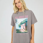 A woman wearing an oversized gray graphic t-shirt with pink text reading 'ALL ABOUT EVE' and a scenic print, paired with blue jeans, against a light gray studio background.