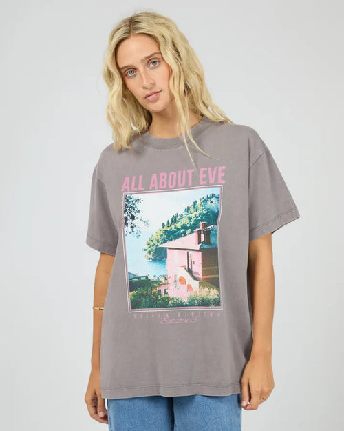 A woman wearing an oversized gray graphic t-shirt with pink text reading 'ALL ABOUT EVE' and a scenic print, paired with blue jeans, against a light gray studio background.