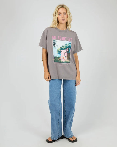 A woman modeling an oversized gray graphic t-shirt with a pink and multicolor print on the front, paired with wide-leg blue jeans and black sandals, against a light gray studio background.