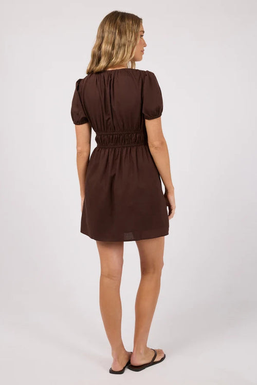 A woman viewed from the back wearing a short brown dress with puff sleeves and a gathered waist, standing against a light gray studio background.