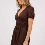 A woman modeling a dark brown short-sleeve mini dress with a deep V-neckline, puff sleeves, and a gathered elastic waist, set against a plain light gray studio background.