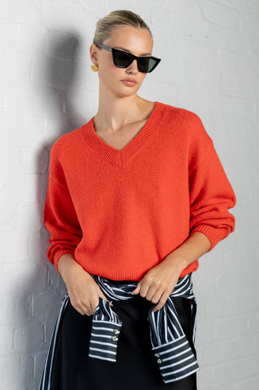A woman wearing a bright orange-red V-neck knit sweater, black sunglasses, and a black skirt with a navy and white striped shirt tied at the waist, posing against a white brick wall.