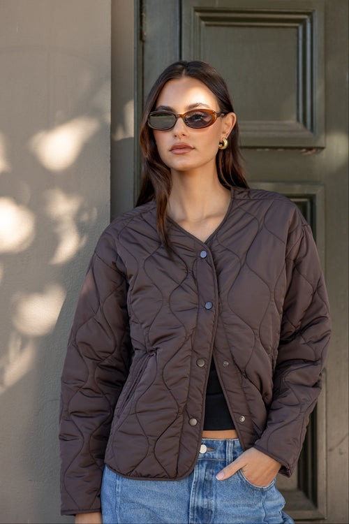 Woman wearing a brown quilted jacket and sunglasses standing against a neutral wall.