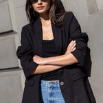 Woman wearing a black blazer and blue jeans standing against a gray wall.