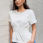 Woman wearing a white top and blue jeans leaning against a light gray brick wall.