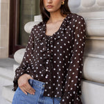 Woman in a polka dot blouse and jeans leaning against a building.