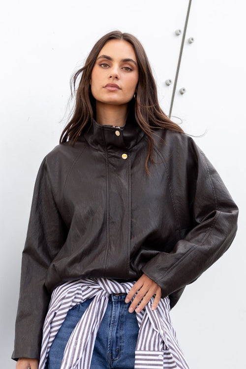Woman wearing a dark jacket, striped shirt, and blue jeans against a white wall.
