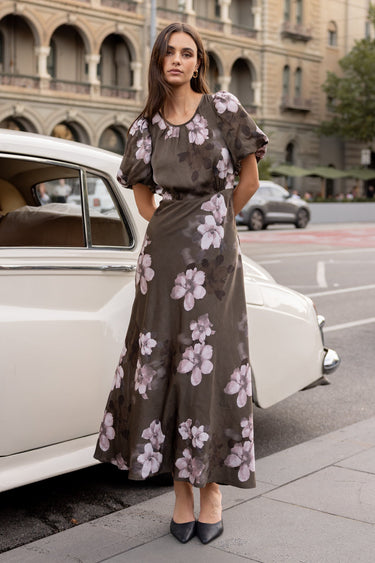 Woman in a floral dress standing next to a vintage car on a city street.
