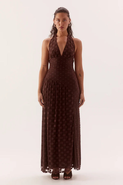 A woman modeling a dark brown halter-neck maxi dress with a subtle polka dot pattern, standing against a plain off-white studio background.
