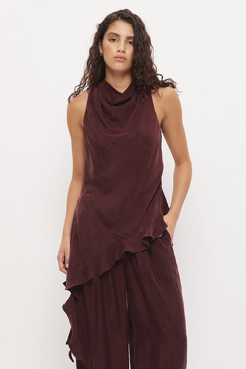 A woman modeling a sleeveless, dark burgundy draped top and matching wide-leg pants set, paired with black flip-flops, against a plain white studio background.