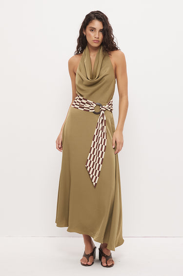 A woman modeling a sleeveless olive green halter maxi dress with a cowl neck, a matching tie belt, and a brown and white checkered patterned scarf tied at the waist, set against a plain white studio background.