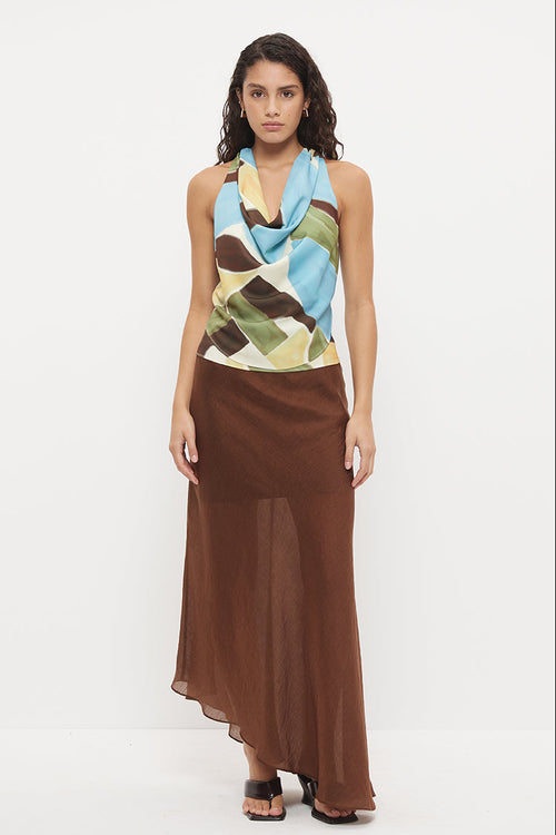 A model stands against a plain white studio background wearing a sleeveless halter-neck maxi dress with a multicolor abstract print on the bodice and a flowing brown skirt.