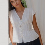 Woman wearing a white blouse and dark pants standing in front of a white curtain.