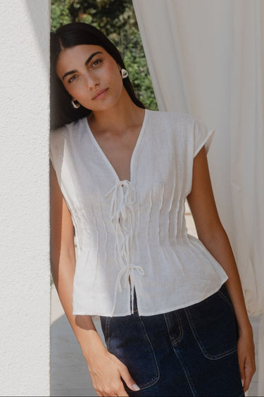 Woman wearing a white blouse and dark pants standing in front of a white curtain.