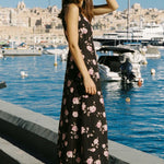 Woman in a floral dress standing by a waterfront with boats and buildings in the background