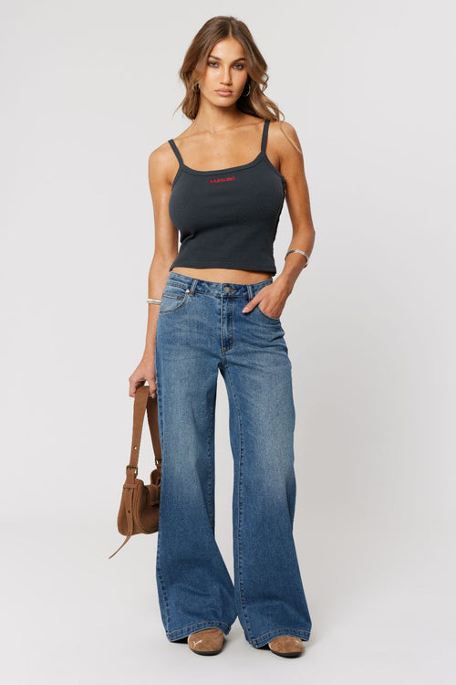 Woman wearing a black tank top and blue jeans holding a brown bag on a white background