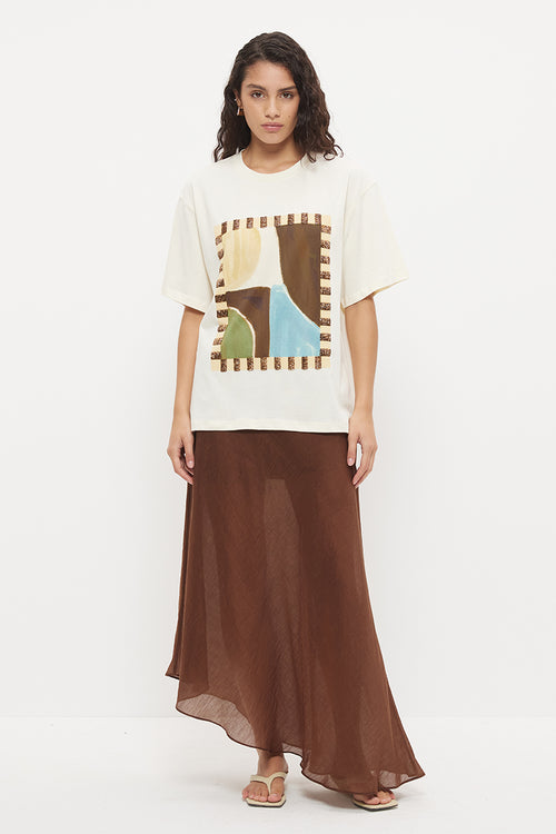A model stands against a plain white studio background wearing a cream-colored oversized graphic t-shirt with a brown abstract print and a long brown skirt.