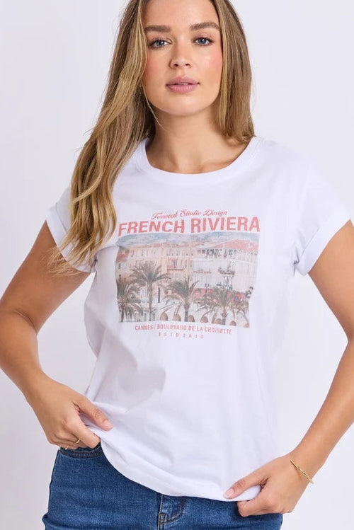 A woman modeling a white short-sleeve graphic T-shirt with a red and beige 'French Riviera' print on the front, paired with blue jeans against a plain white background.