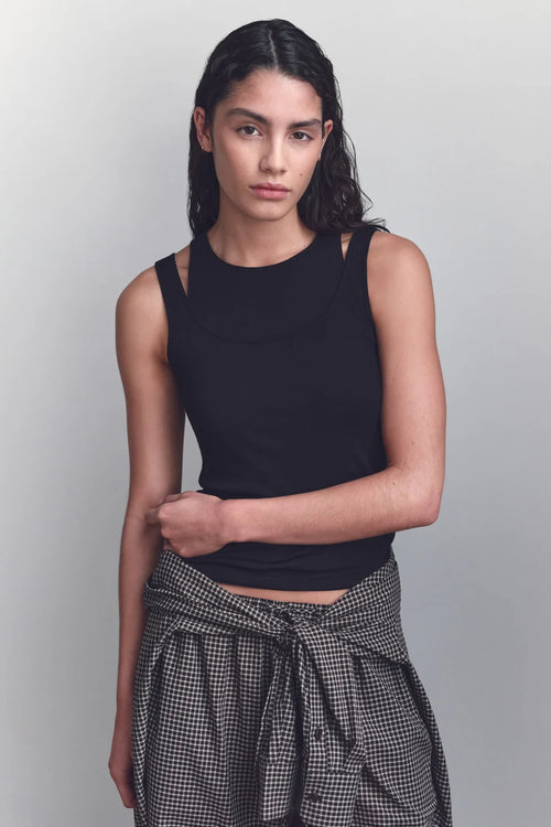 A woman modeling a black sleeveless crop top with side cutouts and a high neckline, paired with a black and white gingham shirt tied around her waist, against a light gray studio background.