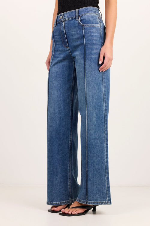 A model wearing a pair of wide-leg blue denim jeans with a black top and black flat sandals, shown from the waist down against a plain white studio background.