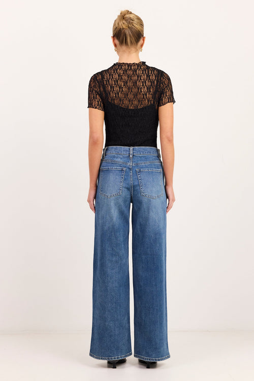 A woman viewed from the back wearing a black short-sleeve lace-trim top and blue wide-leg jeans against a plain light gray studio background.