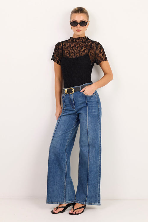 A model stands against a plain white studio background wearing a black short-sleeve top with a sheer lace upper section, blue wide-leg jeans with a gold belt, and black sandals.