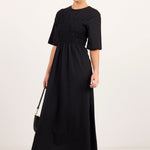 A woman modeling a black midi dress with short sleeves, a round neckline, and a smocked waist, styled with black sunglasses, black flats, and a small black handbag against a plain white studio background.